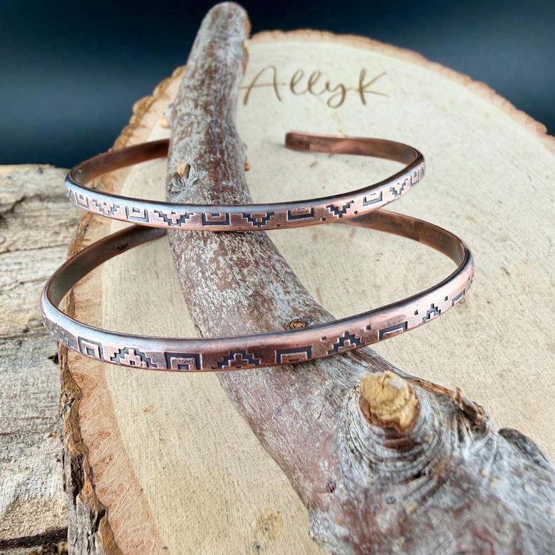 May include: Two thin copper cuff bracelets featuring a dark, etched geometric pattern of repeating squares and stepped shapes. The bracelets are open-ended and display a warm, metallic finish with oxidised details within the engraved designs.