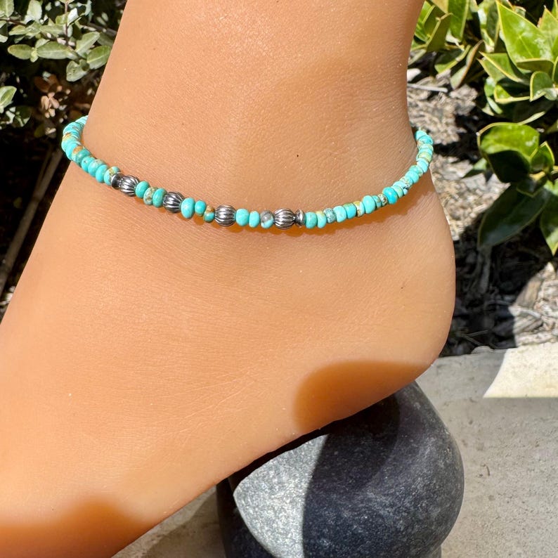 May include: Turquoise beaded anklet with silver-colored accents. The anklet features small, round turquoise beads interspersed with larger, textured silver beads. The jewelry is designed to be worn around the ankle.