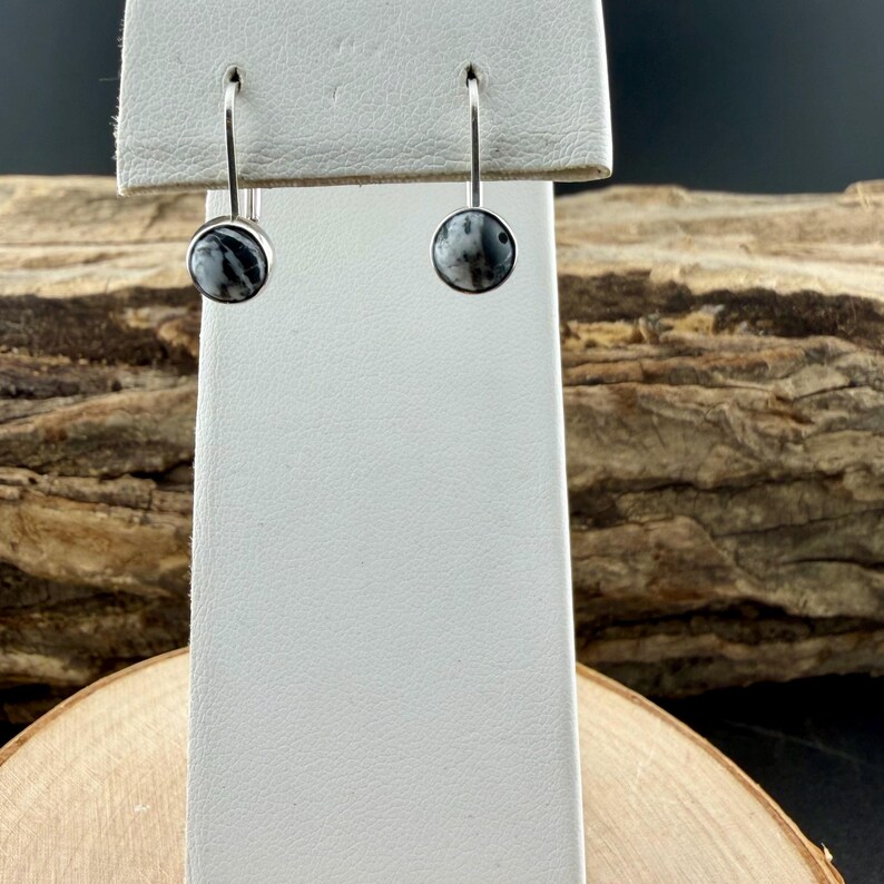 May include: A pair of silver drop earrings. Each earring features a round, marbled stone with black and white patterns. The earrings are displayed on a white earring stand against a natural wood background.