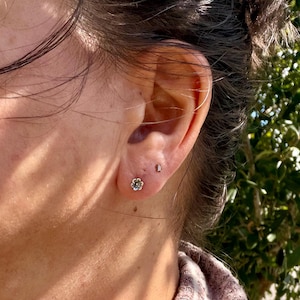 May include: A silver-toned stud earring shaped like a small flower with five petals and a central dot, positioned on an earlobe next to a tiny, slender, rectangular-shaped silver-toned stud earring located slightly higher up.