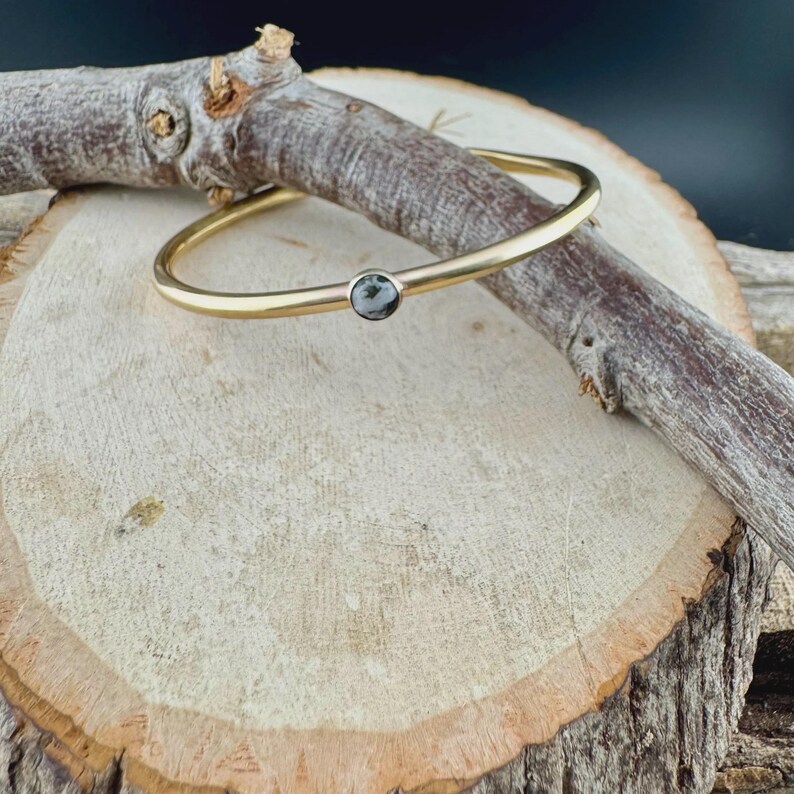 May include: A gold-colored bangle bracelet with a small, round, gray and white stone set in the center. The bracelet is resting on a piece of wood with a natural, light brown color and a dark brown branch.