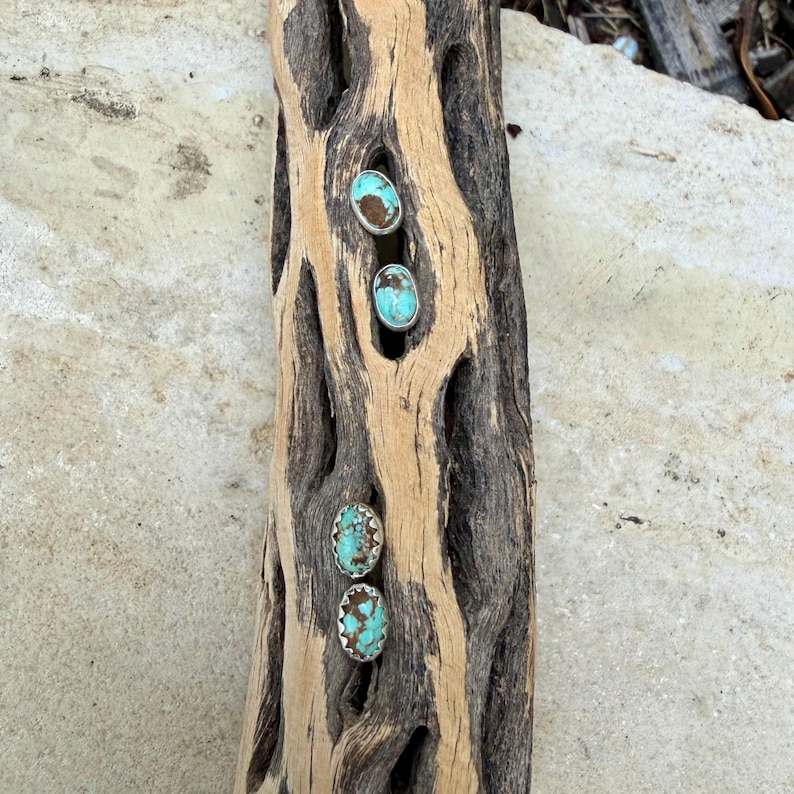 May include: Four pairs of turquoise and silver earrings are displayed on a piece of weathered wood. The earrings have a bright turquoise stone set in a silver bezel. The wood has a natural, light brown color with dark brown holes.