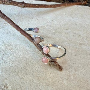 May include: Two silver rings featuring round, smooth, light pink cabochon stones. Each ring has an open-ended, bypass-style band with a hammered texture. The rings are positioned on a thin, brown wooden twig, showcasing the metallic finish.