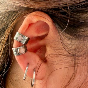 May include: Two silver ear cuffs featuring textured metal bands and round, light pink cabochon stones. Below these, two silver hoop earrings are visible, one with a twisted rope design and the other a smooth, thin, circular metal band.