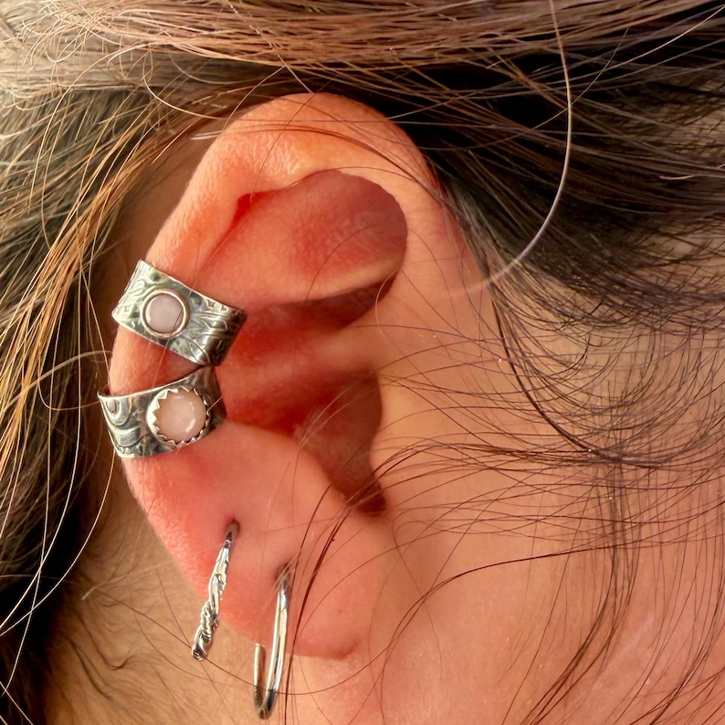 May include: Two silver ear cuffs featuring textured metal bands and round, light pink cabochon stones. Below these, two silver hoop earrings are visible, one with a twisted rope design and the other a smooth, thin, circular metal band.