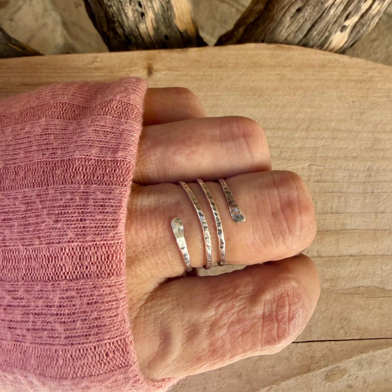 May include: A silver-toned bypass ring featuring a textured, hammered surface. The ring is designed with three distinct, overlapping bands that wrap around the finger, creating a layered appearance with a metallic, reflective finish.
