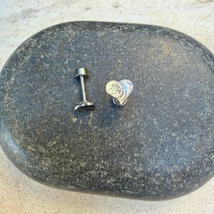 May include: A silver-toned metal labret stud featuring a flat heart-shaped top with an embossed, swirling line pattern. The jewelry piece consists of a straight post with a circular base and a detachable heart-shaped front component.