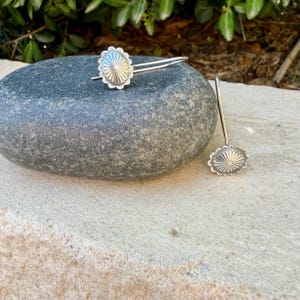 May include: A pair of silver-toned earrings featuring a circular, sunburst-patterned design with scalloped edges. Each earring has a thin, curved wire hook for attachment, showcasing a metallic finish with intricate radial lines radiating from the center.