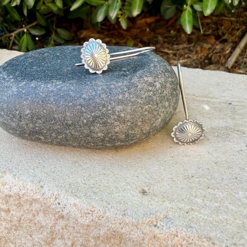 May include: A pair of silver-toned earrings featuring a circular, sunburst-patterned design with scalloped edges. Each earring has a thin, curved wire hook for attachment, showcasing a metallic finish with intricate radial lines radiating from the center.