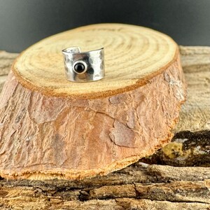 May include: A silver-toned ring with a hammered texture and a small, round, black stone. The ring is displayed on a wooden surface with a natural, rustic appearance. The ring is a piece of jewelry.