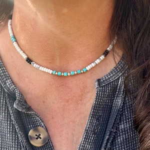 May include: A beaded necklace with white, black, turquoise, and gold-colored beads. The necklace is worn around the neck and is made of small, round beads. The turquoise beads are square-shaped and interspersed with gold-colored beads.