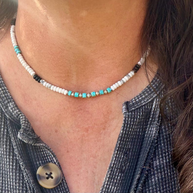 May include: A beaded necklace with white, black, turquoise, and gold-colored beads. The necklace is worn around the neck and is made of small, round beads. The turquoise beads are square-shaped and interspersed with gold-colored beads.