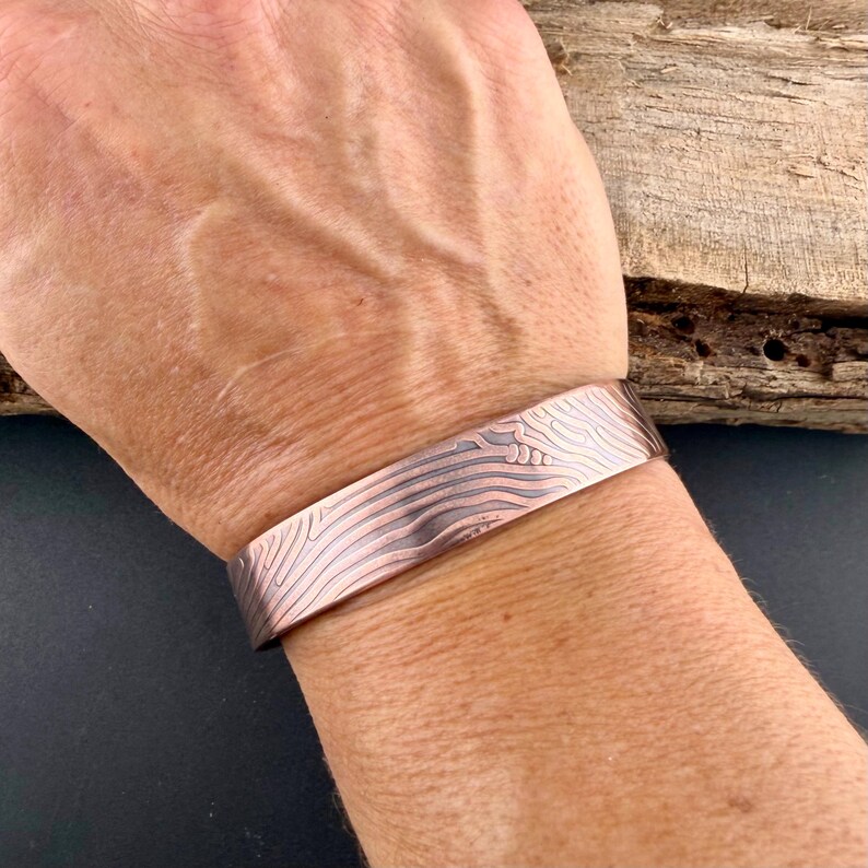 May include: A copper cuff bracelet with a textured, wave-like pattern. The bracelet is wide and wraps around the wrist. The copper has a reddish-brown hue with lighter, silver-colored lines creating the design. The bracelet is a piece of jewelry.