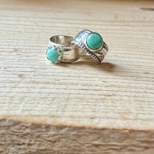 May include: Two silver rings featuring round turquoise stones. The first ring has a smooth band with decorative cutouts, while the second ring displays an engraved pattern along the band. Both rings are set with vibrant blue-green stones.