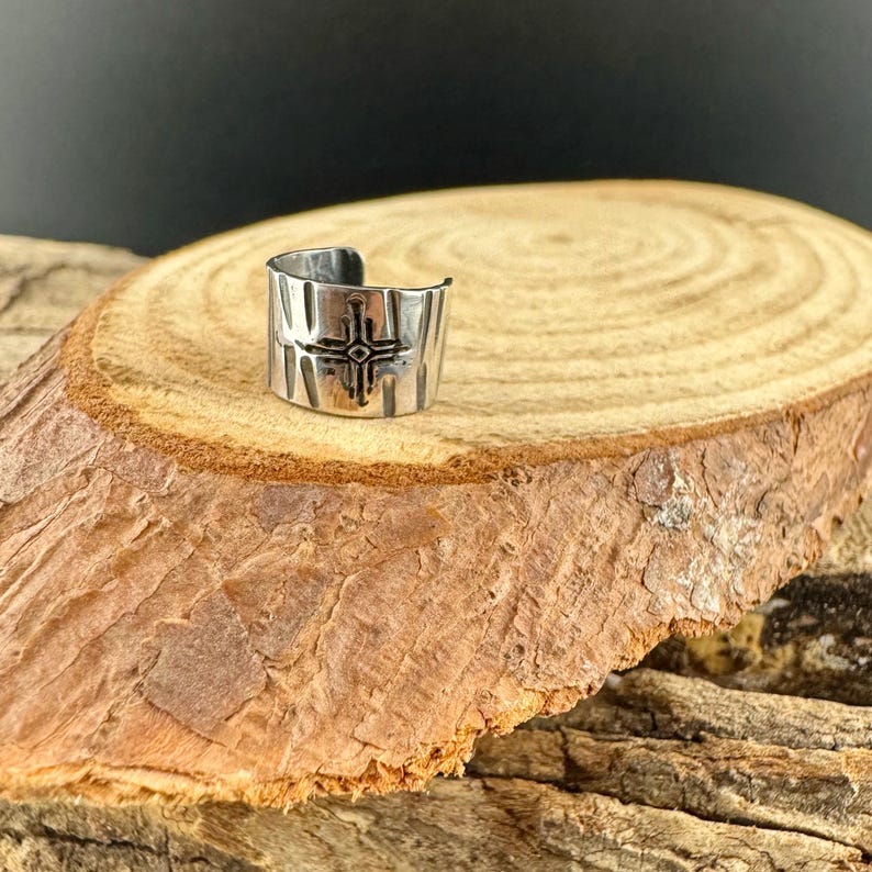 Puede incluir: Un anillo plateado con un dise&ntilde;o de cruz geom&eacute;trica. El anillo es ancho y tiene una superficie texturizada. Se muestra sobre una superficie de madera con un aspecto natural y r&uacute;stico.