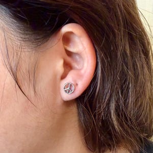 Puede incluir: Primer plano de un pendiente de bot&oacute;n con el s&iacute;mbolo de la paz plateado en una oreja. El pendiente es un dise&ntilde;o peque&ntilde;o y circular con el s&iacute;mbolo de la paz en el centro. El pelo de la persona es casta&ntilde;o.