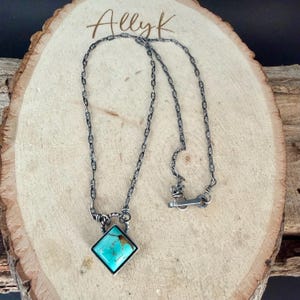 May include: A silver-toned necklace with a turquoise gemstone pendant. The pendant is a square-shaped stone with a silver border. The necklace is on a wooden surface with the name "Allyk" written in cursive.