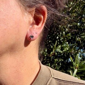 May include: A small, silver-toned heart-shaped stud earring worn in the earlobe. The earring features a polished, reflective surface and a simple, compact design that sits flush against the skin, catching the natural sunlight.