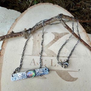 May include: A silver necklace featuring a rectangular bar pendant with an intricate embossed pattern and a central oval iridescent stone. The pendant is attached to a delicate silver chain with a lobster claw clasp closure.