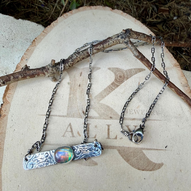 May include: A silver necklace featuring a rectangular bar pendant with an intricate embossed pattern and a central oval iridescent stone. The pendant is attached to a delicate silver chain with a lobster claw clasp closure.
