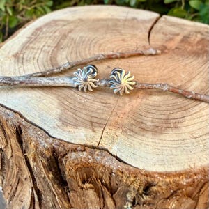 May include: A pair of silver-toned stud earrings featuring a sunburst design with radiating rays. The earrings are resting on a thin, light brown twig that is positioned across the center of a circular, light-colored wooden log slice.