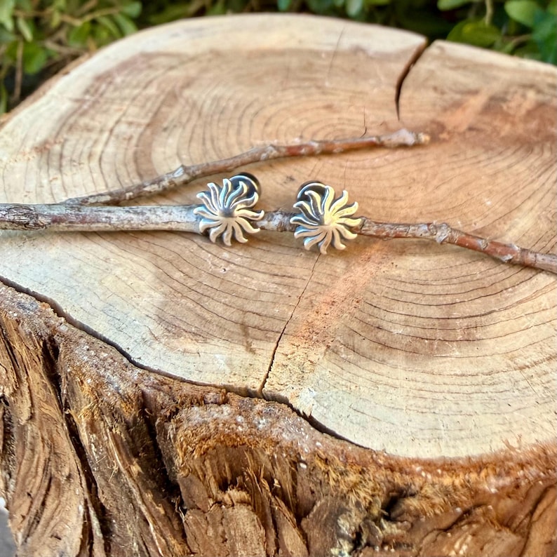 May include: A pair of silver-toned stud earrings featuring a sunburst design with radiating rays. The earrings are resting on a thin, light brown twig that is positioned across the center of a circular, light-colored wooden log slice.