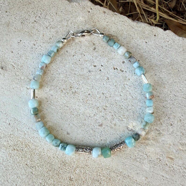 Puede incluir: Una pulsera de cuentas con peque&ntilde;as piedras de corte cuadrado en azul claro y verde p&aacute;lido, intercaladas con espaciadores cil&iacute;ndricos de tono plateado. La pulsera se asegura con un cierre de mosquet&oacute;n plateado y una peque&ntilde;a argolla.
