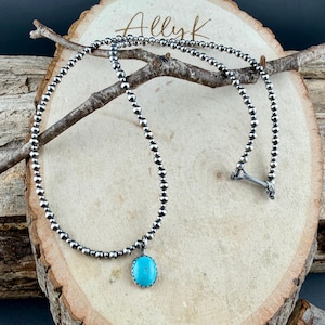 May include: A silver beaded necklace with a turquoise pendant. The necklace is displayed on a wooden surface with a branch. The pendant is an oval-shaped turquoise stone with a silver bezel setting. The necklace has a bone-shaped clasp.