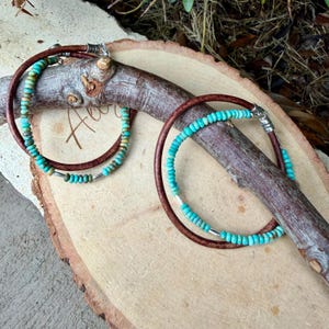May include: Two circular bracelets featuring brown leather cords adorned with small, round, turquoise-colored beads and interspersed with thin, cylindrical silver-toned metal spacers. Each bracelet is secured with a silver-toned metal clasp.