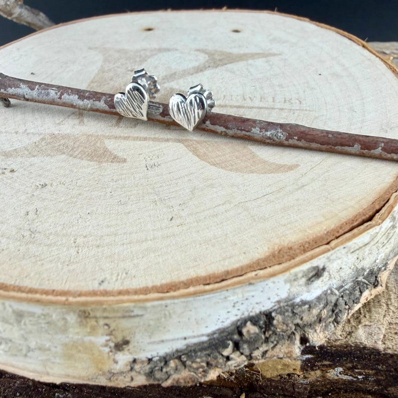 May include: A pair of silver heart-shaped stud earrings with a textured, brushed finish. The earrings are positioned on a thin, horizontal wooden twig, showcasing the metallic luster and the detailed surface pattern of the heart design.