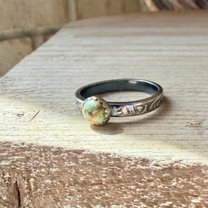 May include: A silver ring with a turquoise stone. The ring has a dark band and a decorative pattern around the outside. The turquoise stone is oval-shaped and set in a starburst setting.