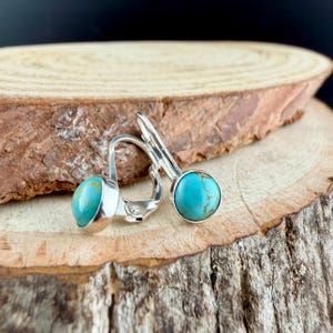 May include: Silver leverback earrings featuring round, turquoise stones. The earrings are presented on a wooden surface, highlighting the bright blue-green hue of the stones. The design is simple and elegant, suitable for everyday wear.