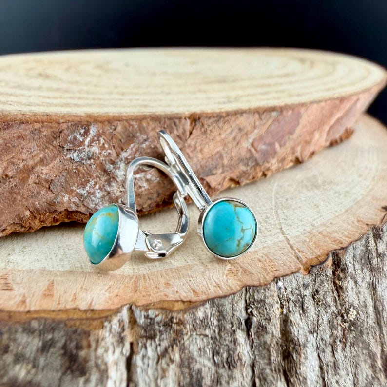 May include: Silver leverback earrings featuring round, turquoise stones. The earrings are presented on a wooden surface, highlighting the bright blue-green hue of the stones. The design is simple and elegant, suitable for everyday wear.