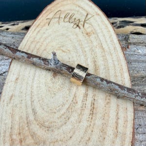May include: A gold-colored ring with a textured surface, resting on a wooden branch. The ring has a wide band design. The background features a wooden slice with the handwritten text "Allyk".