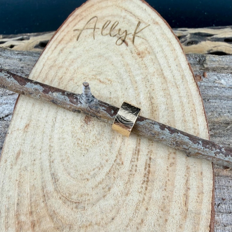 May include: A gold-colored ring with a textured surface, resting on a wooden branch. The ring has a wide band design. The background features a wooden slice with the handwritten text "Allyk".