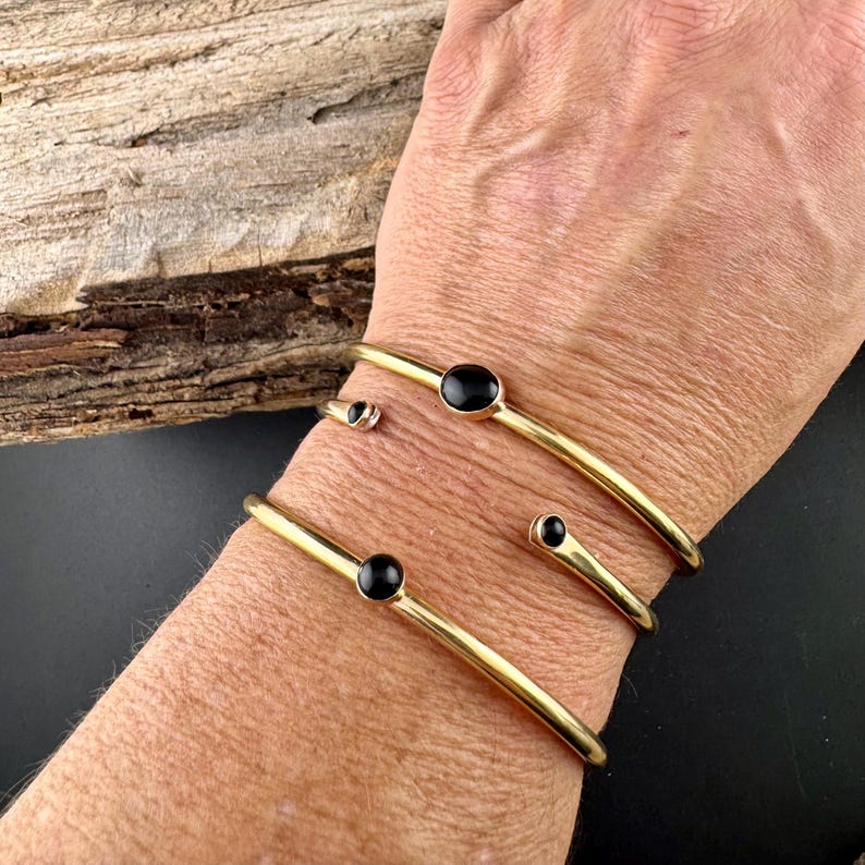 May include: Gold-toned metal bracelets with black stone accents. The bracelets are worn on a wrist, with a dark background. The bracelets have a minimalist design, with a smooth, polished finish.