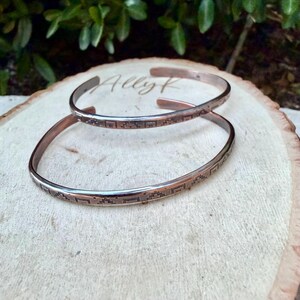 May include: Two thin copper cuff bracelets featuring a repeating geometric pattern etched along the outer surface. The bracelets are open-ended and have a metallic finish with dark oxidised detailing within the engraved square and step designs.