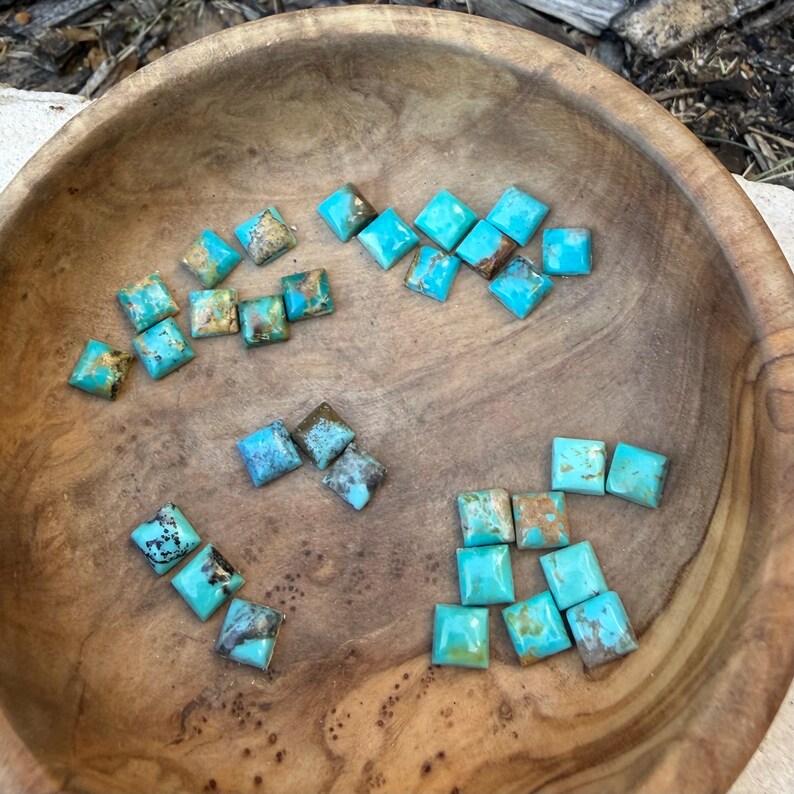Op de afbeelding: Een houten kom gevuld met vierkante turkoois edelstenen. De edelstenen hebben een levendige turkooiskleur met bruine en zwarte matrixpatronen. De kom heeft een natuurlijke houtnerf afwerking.
