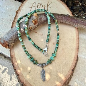 May include: A turquoise beaded necklace with a silver feather pendant. The necklace is displayed on a wooden surface. The text "Allyk" is written in cursive on the wood.