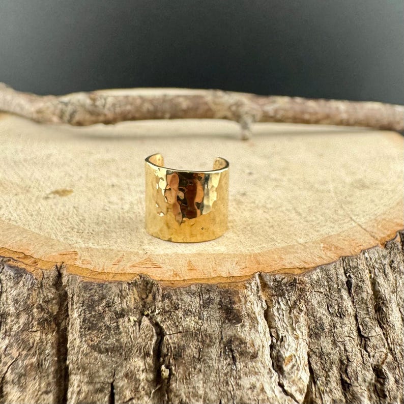May include: A gold ring with a hammered texture. The ring is a wide band style, with a slightly open design. The ring is displayed on a wooden surface, with a dark background.