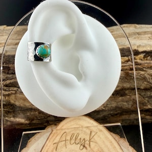 May include: A silver ear cuff with a hammered texture and a round turquoise stone. The cuff is displayed on a white ear-shaped form. The jewelry is presented on a wooden surface.