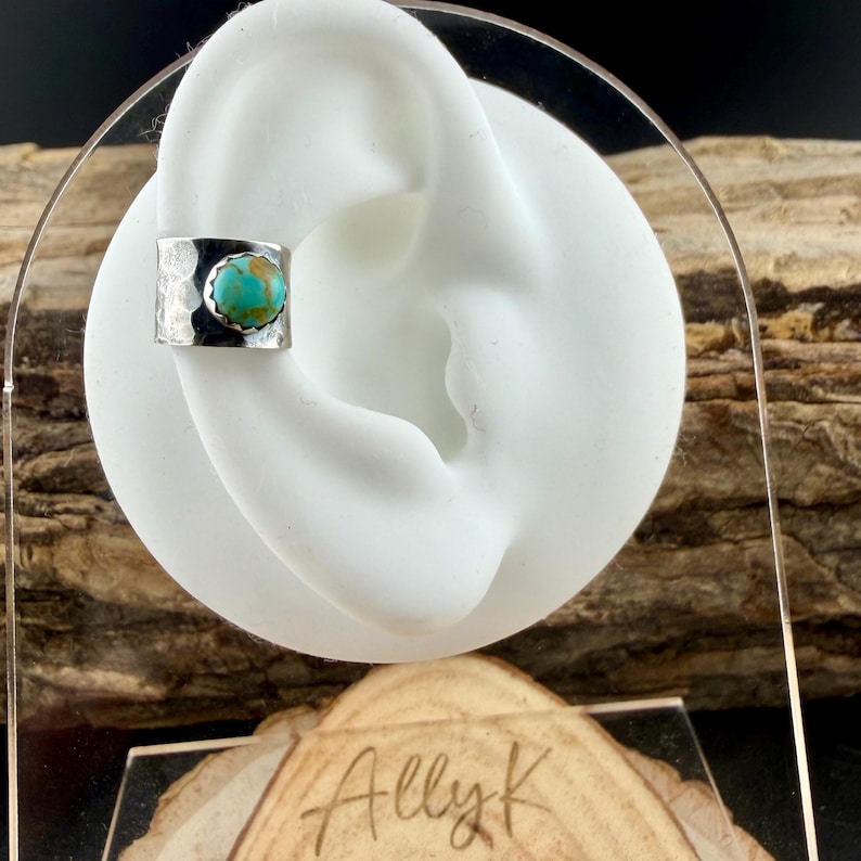 May include: A silver ear cuff with a hammered texture and a round turquoise stone. The cuff is displayed on a white ear-shaped form. The jewelry is presented on a wooden surface.
