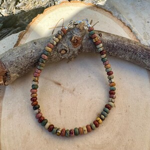 May include: A beaded bracelet featuring a variety of earth-toned beads in shades of brown, green, red, and yellow. The beads are strung together with a silver-colored clasp. The bracelet is displayed on a wooden surface.