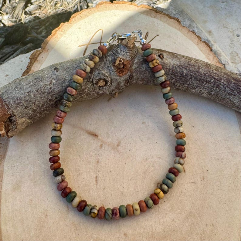 May include: A beaded bracelet featuring a variety of earth-toned beads in shades of brown, green, red, and yellow. The beads are strung together with a silver-colored clasp. The bracelet is displayed on a wooden surface.