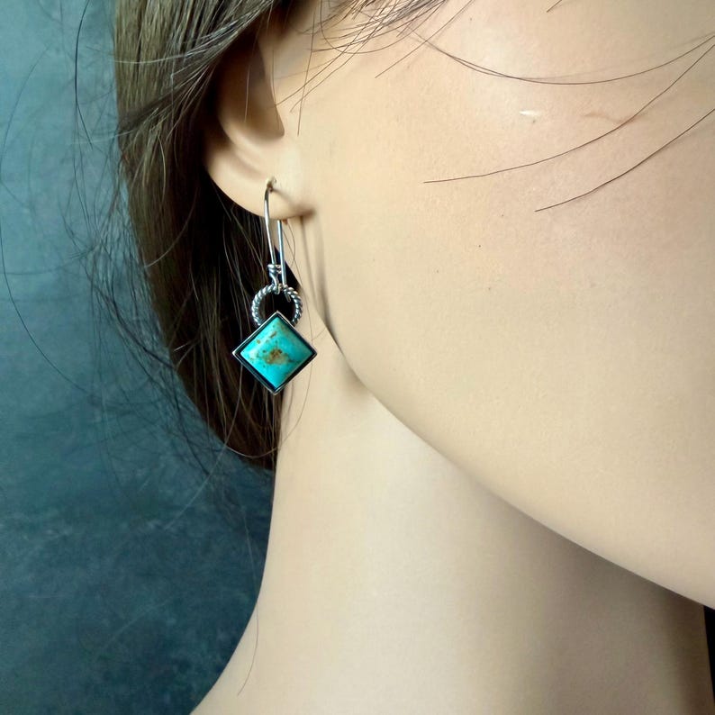 May include: Silver-toned dangle earrings featuring a square turquoise stone. The turquoise stone is set in a silver frame and hangs from a decorative silver loop. The earrings are shown on a person's ear.