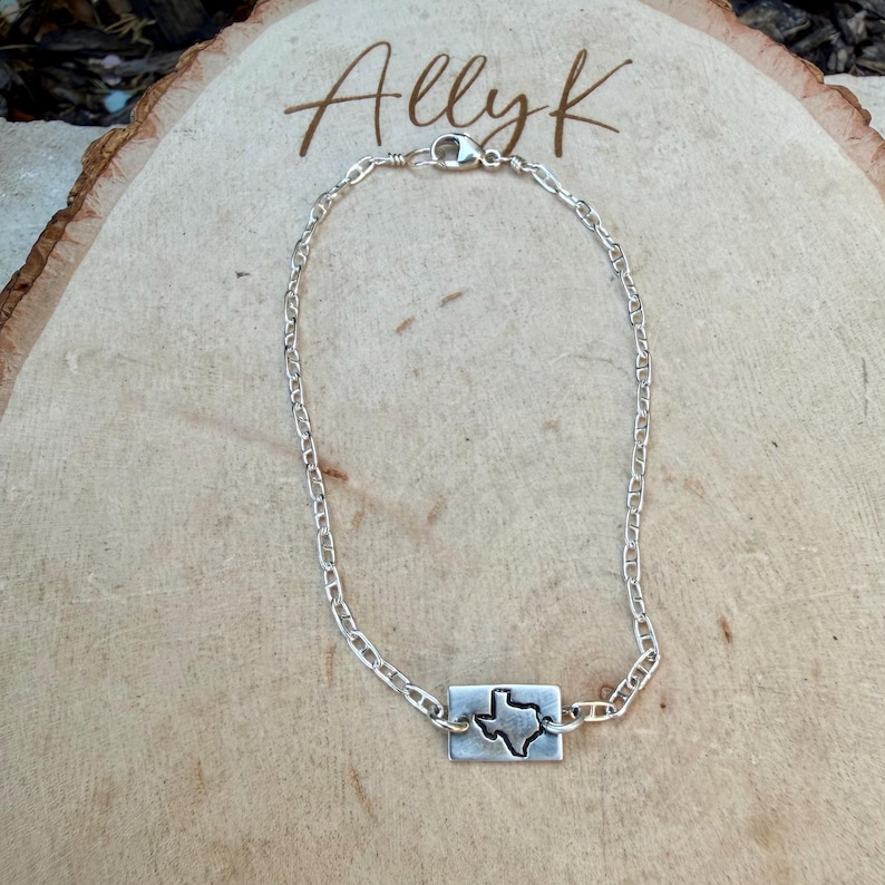 May include: A silver chain bracelet with a rectangular charm featuring a cutout of the state of Texas. The bracelet is displayed on a wooden surface, with the name "Allyk" written in cursive script.