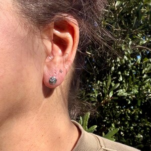 May include: A small, round, silver-toned stud earring featuring a textured, geometric pattern with a central dot. The earring is worn in the lower earlobe, positioned below a tiny, clear, circular stud earring in the upper lobe.