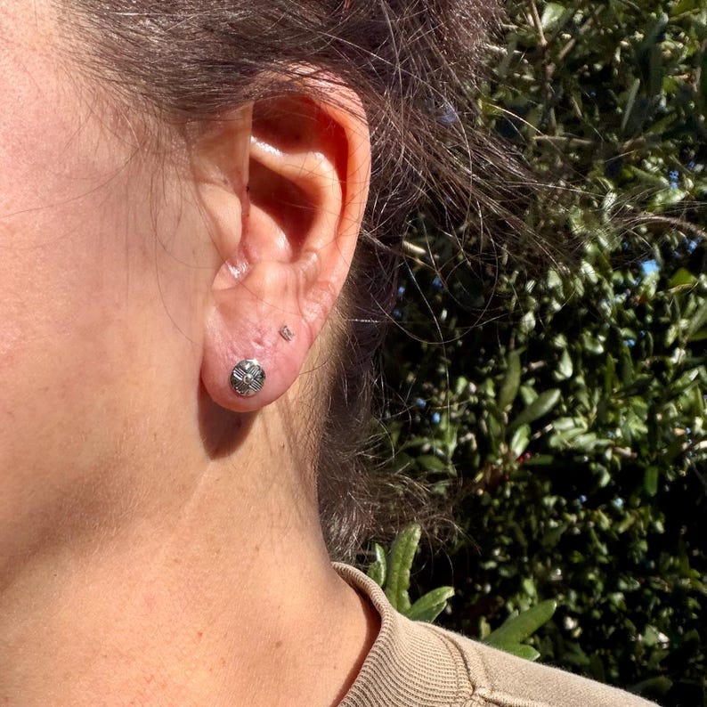 May include: A small, round, silver-toned stud earring featuring a textured, geometric pattern with a central dot. The earring is worn in the lower earlobe, positioned below a tiny, clear, circular stud earring in the upper lobe.