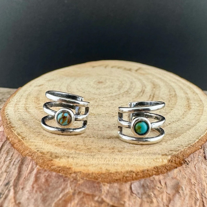May include: Two silver-toned ear cuffs with a layered design, each featuring a round turquoise stone. The ear cuffs are displayed on a wooden surface, showcasing their intricate details and vibrant color.