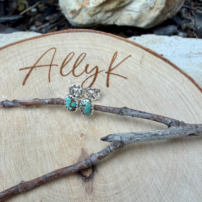 May include: A pair of turquoise and silver stud earrings displayed on a wooden surface with the handwritten text "Ally k". The earrings have a silver border and a turquoise center. The earrings are placed on a small twig.